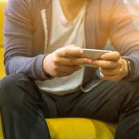 a man wearing a hoodie and a smartwatch, holds a smartphone with both hands while sitting on a yellow sofa engaging in mental telehealth