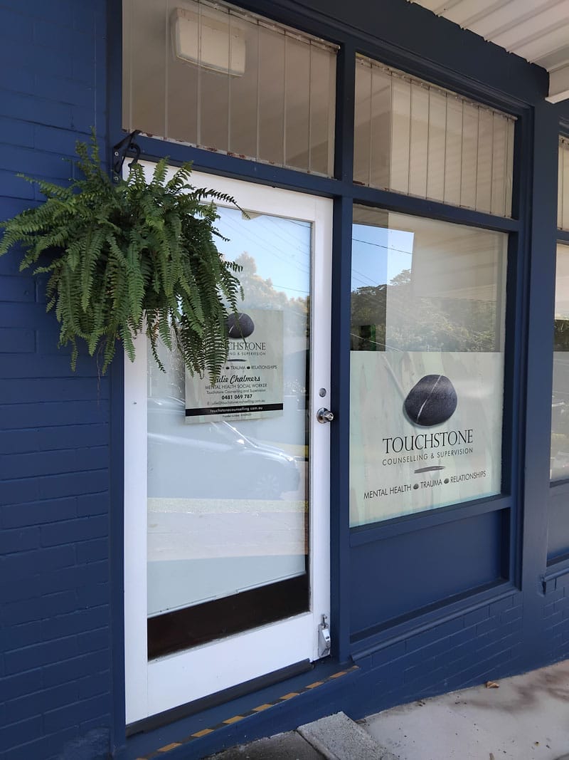 The entrance to Touchstone Counseling & Support, featuring a glass door with a sign, a potted fern hanging above, and an inviting atmosphere that welcomes you to Work With Us.